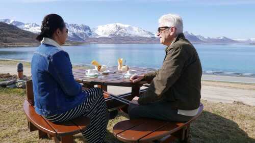 Two persons sitting by a outdoor table looking at the fjord and the mountains